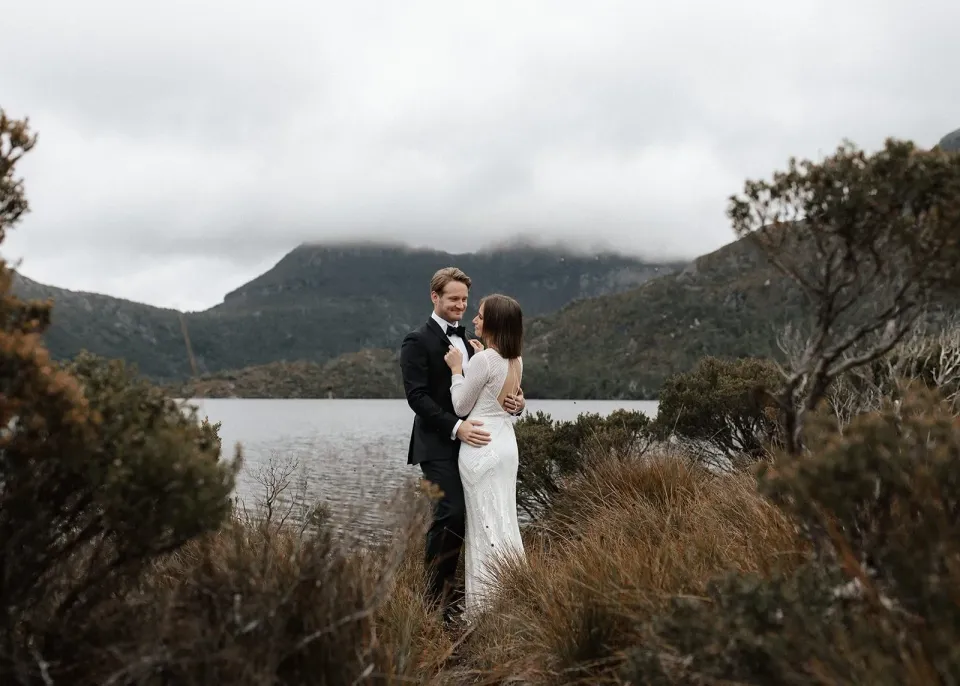 April and Josh standing together among lakeside shrubs with mountains behind them, at Dove Lake, Cradle Mountain, Tasmania, for an Elopement Collective elopement. Photo by Bulb Creative.