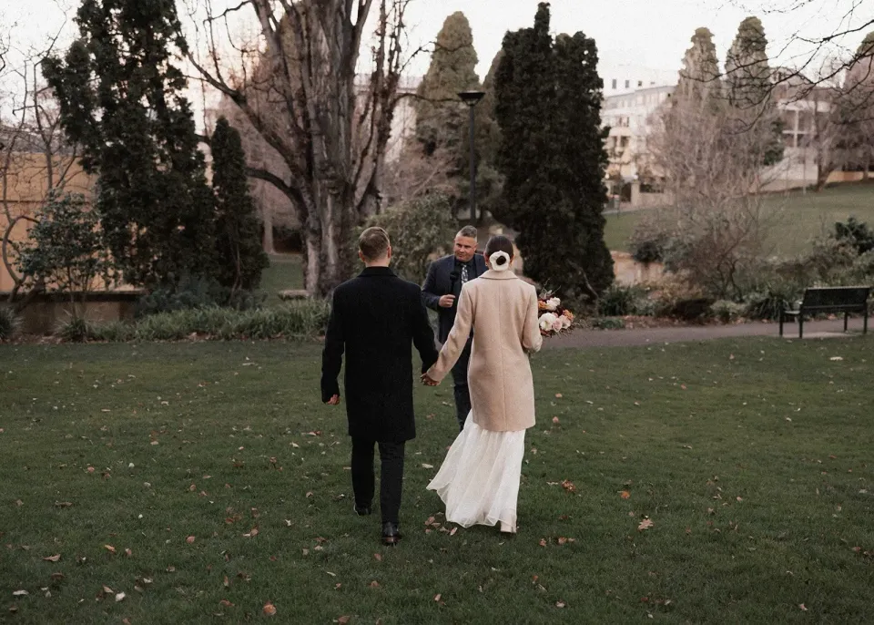 A couple walk hand in hand across a garden lawn towards tall trees.
