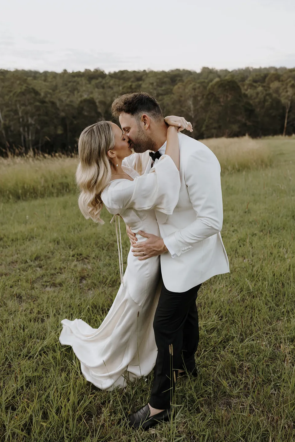 Greg holding Gabrielle in his arms as they kiss in the green paddock, at Roundhill Retreat in Obi Obi, Sunshine Coast Hinterland, Queensland, for an Elopement Collective elopement. Photo by George Bowden.