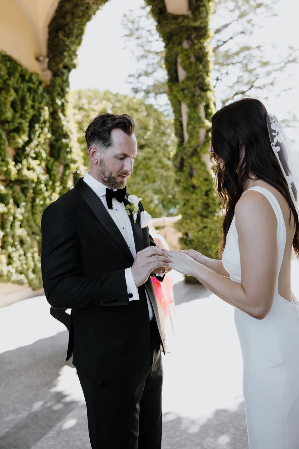 Lachlan placing a wedding ring on Lyndal’s hand beneath Villa del Balbianello’s loggia, with his white calla lily boutonniere by Garden of Love visible, at Lake Como in Lombardy, Italy, for an Elopement Collective elopement. Photograph by George Bowden.