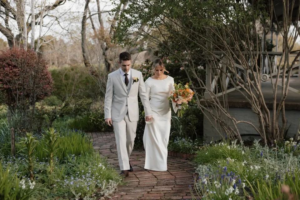 Maddy and Cameron walking hand in hand along a brick garden path, carrying her orange and pink bouquet by The Flower Jar, at The Mayor’s Residence in Daylesford, Victoria, for an Elopement Collective elopement. Photo by Bulb Creative.