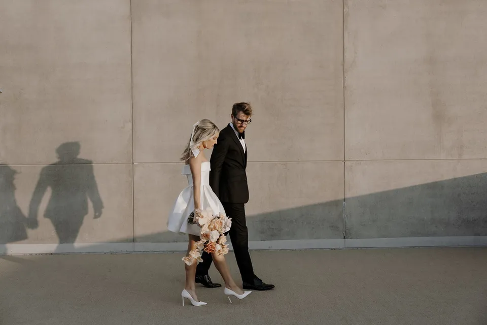 Danielle and Adam walking beside a plain rooftop wall with their shadows falling behind them, at The Calile Hotel in Fortitude Valley, Brisbane, for an Elopement Collective elopement. Photo by George Bowden.
