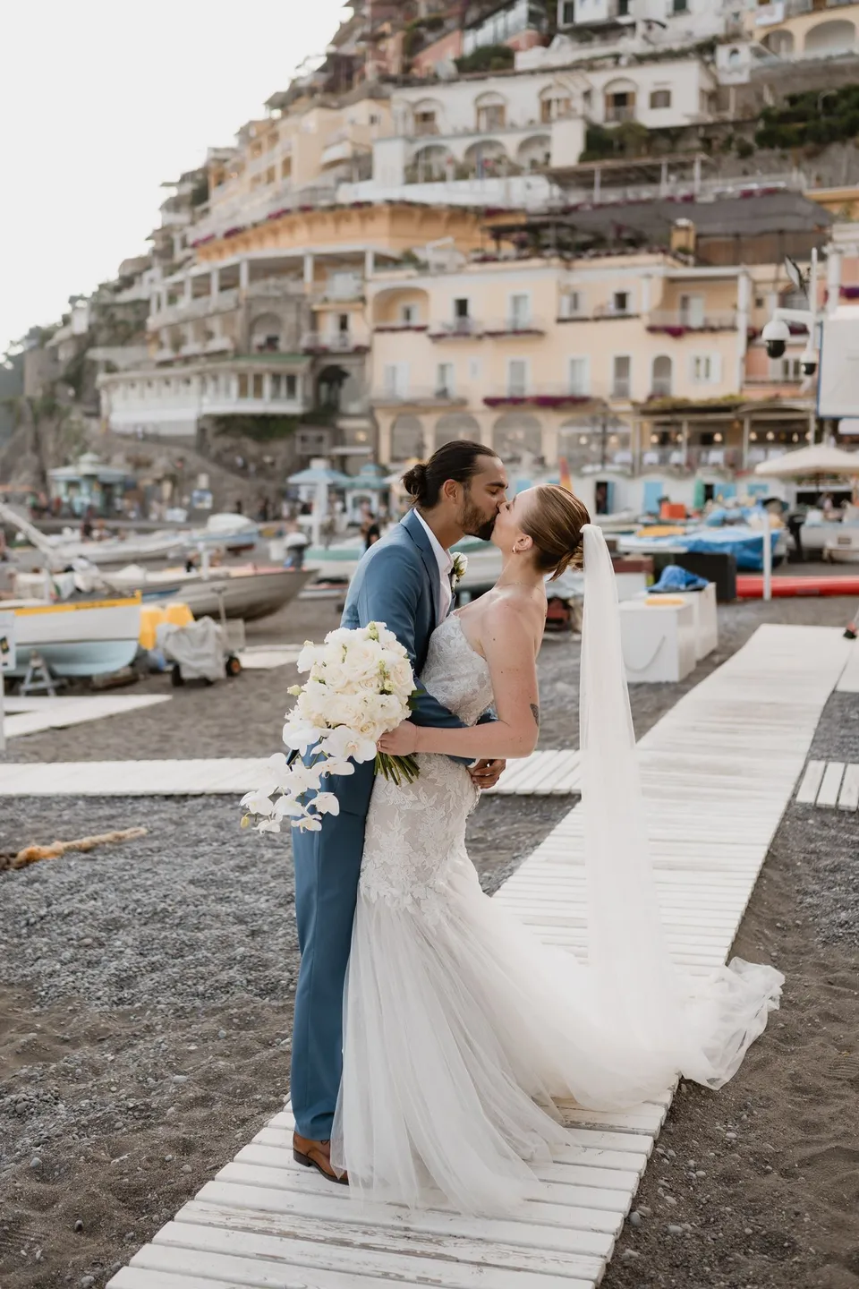 Skyler and Zach kissing on a beach boardwalk with her Flora Garden - Positano bouquet, boats and the hillside town behind them, in Positano on the Amalfi Coast, Italy, for an Elopement Collective elopement. Photograph by Jason Corroto.