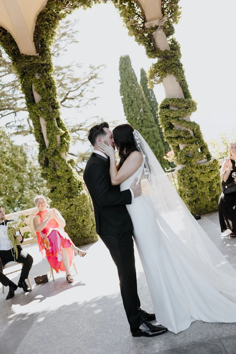 Lyndal and Lachlan kissing during their ceremony beneath Villa del Balbianello’s ivy-covered loggia while their guests look on, at Lake Como in Lombardy, Italy, for an Elopement Collective elopement. Photograph by George Bowden.