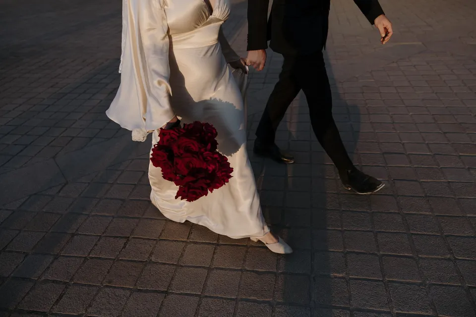 Close view of Kara and Samuel walking hand in hand across sunlit paving, with her white dress, red Ivy Bleu bouquet and his black trousers, at the Louvre in Paris, France, during an Elopement Collective elopement. Photograph by George Bowden.