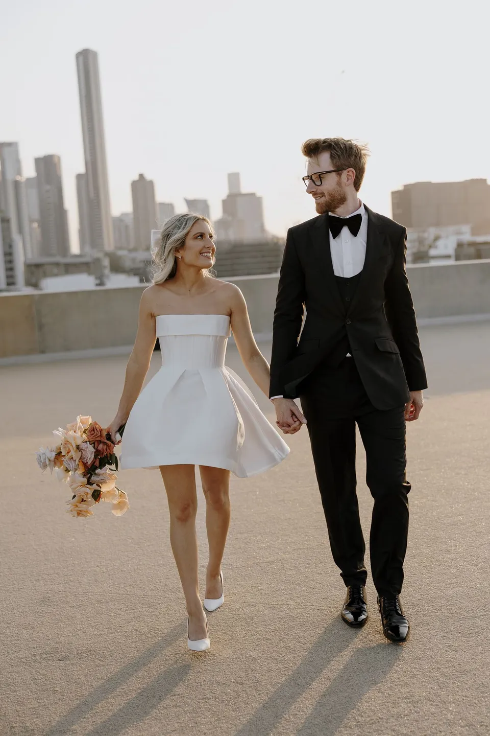 Danielle and Adam looking at each other as they walk hand in hand with Brisbane’s skyline behind them, at The Calile Hotel in Fortitude Valley, Brisbane, for an Elopement Collective elopement. Photo by George Bowden.