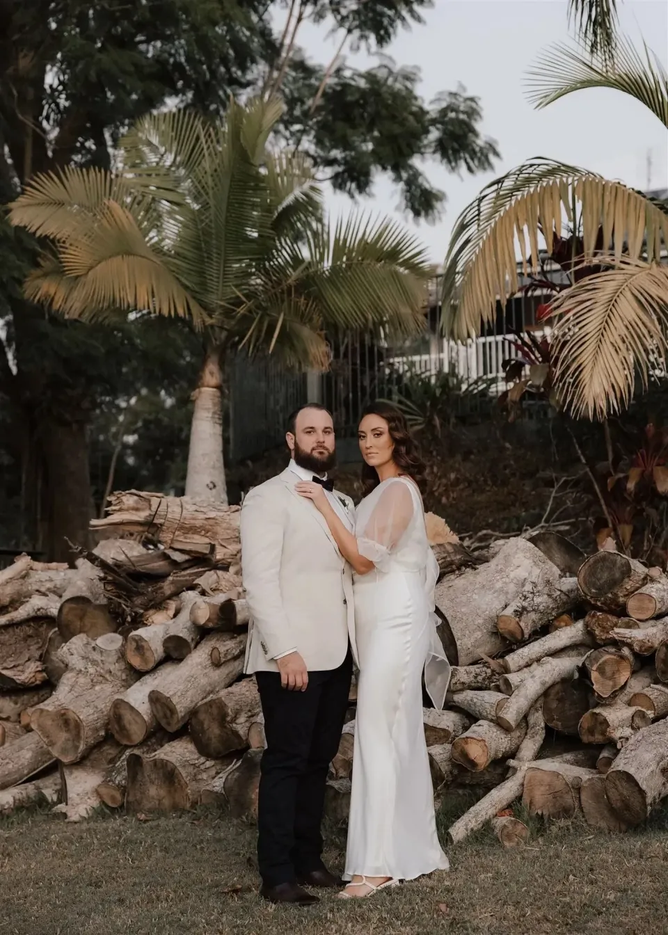 Tineeka and Chris standing together beside a stack of timber logs and tall palms, in Montville, Sunshine Coast Hinterland, Queensland, for an Elopement Collective elopement. Photo by Bulb Creative.