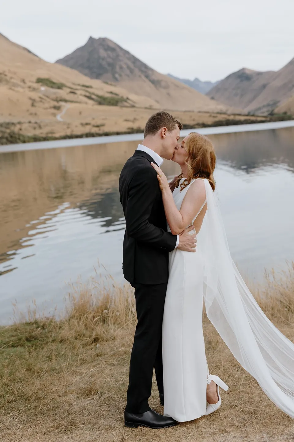 Neve and Hamish kissing after their vows beside the lake, with mountains reflected behind them, at Moke Lake in Queenstown, New Zealand, for an Elopement Collective Queenstown elopement. Photo by George Bowden.