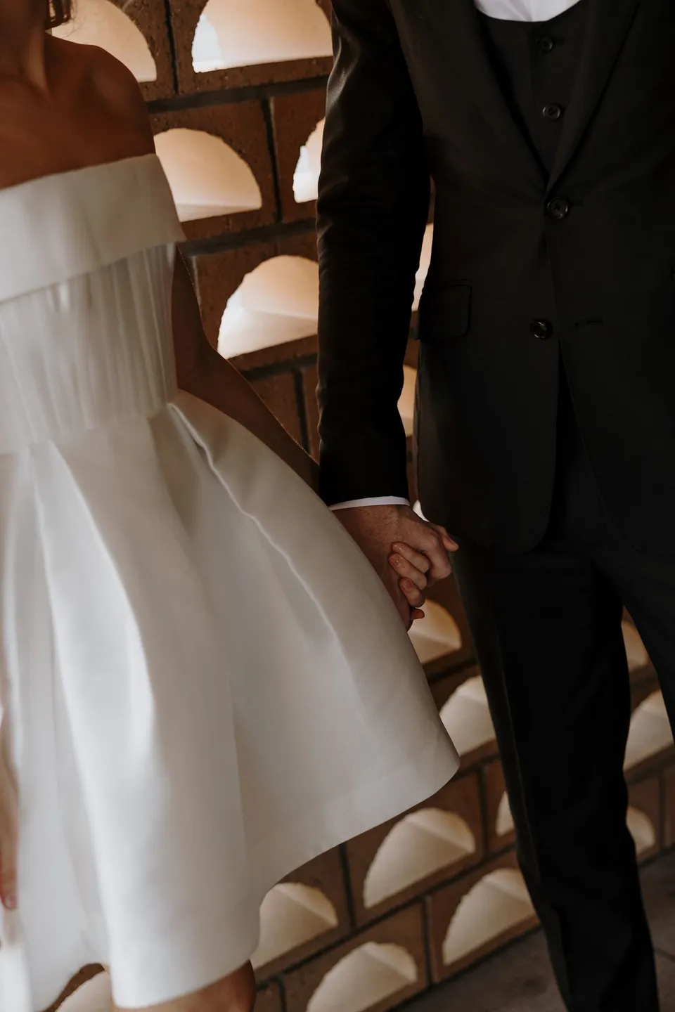 Close view of Danielle and Adam holding hands beside an arched brick screen, at The Calile Hotel in Fortitude Valley, Brisbane, for an Elopement Collective elopement. Photo by George Bowden.
