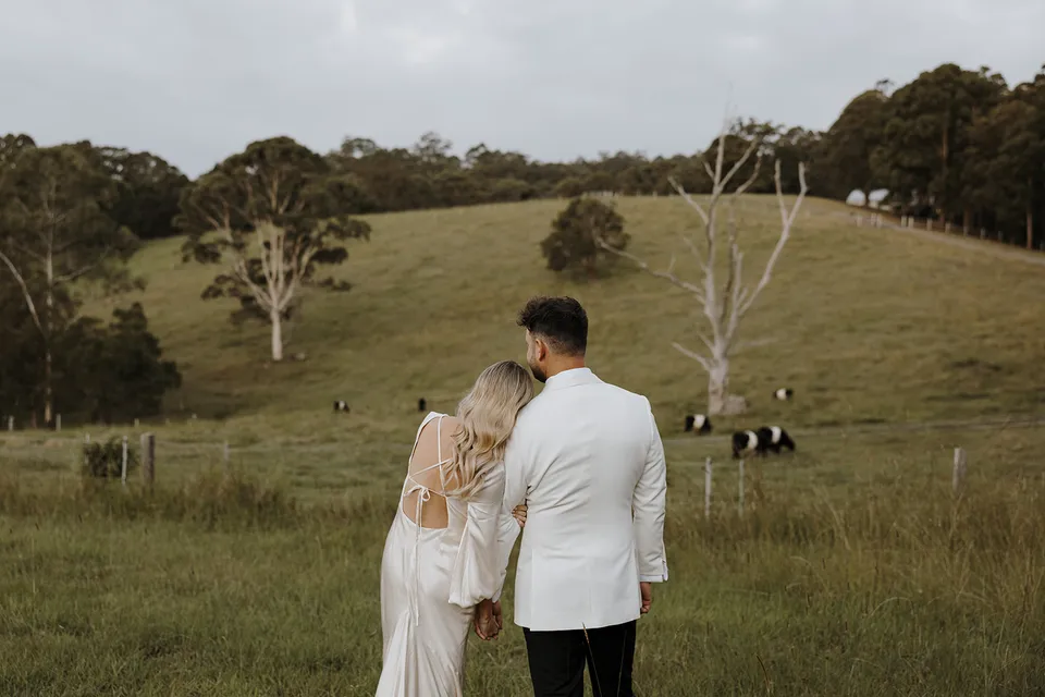 Gabrielle leaning on Greg’s shoulder as they look over rolling green paddocks and grazing cattle, at Roundhill Retreat in Obi Obi, Sunshine Coast Hinterland, Queensland, for an Elopement Collective elopement. Photo by George Bowden.