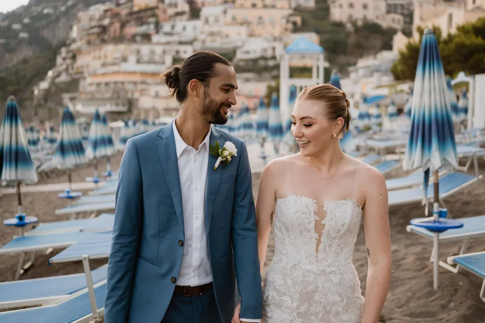 Skyler and Zach smiling together beside blue beach umbrellas, with pastel houses climbing the hillside behind them, in Positano on the Amalfi Coast, Italy, for an Elopement Collective elopement. Photograph by Jason Corroto.