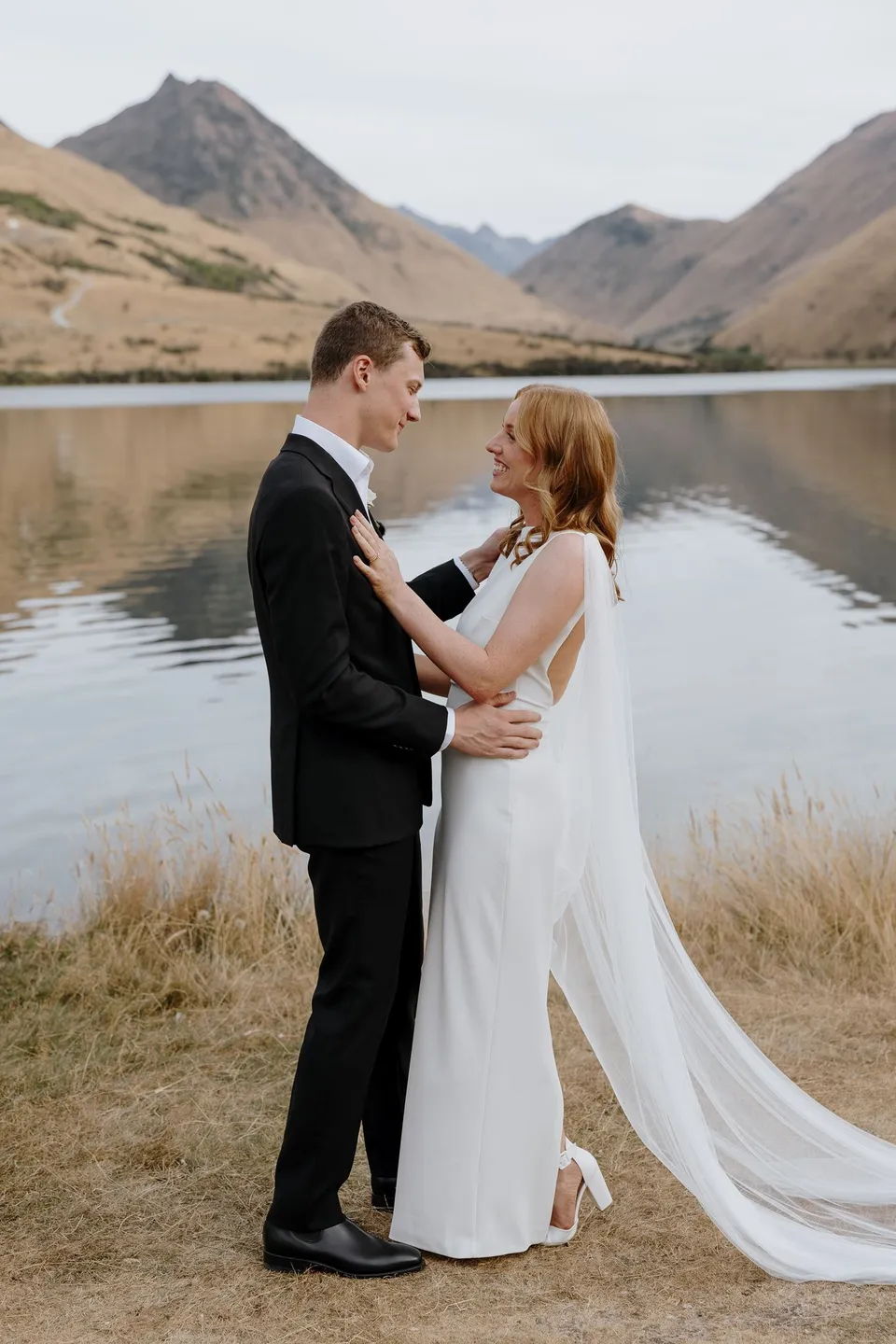 Neve and Hamish smiling and embracing beside the lake after their ceremony, at Moke Lake in Queenstown, New Zealand, for an Elopement Collective Queenstown elopement. Photo by George Bowden.