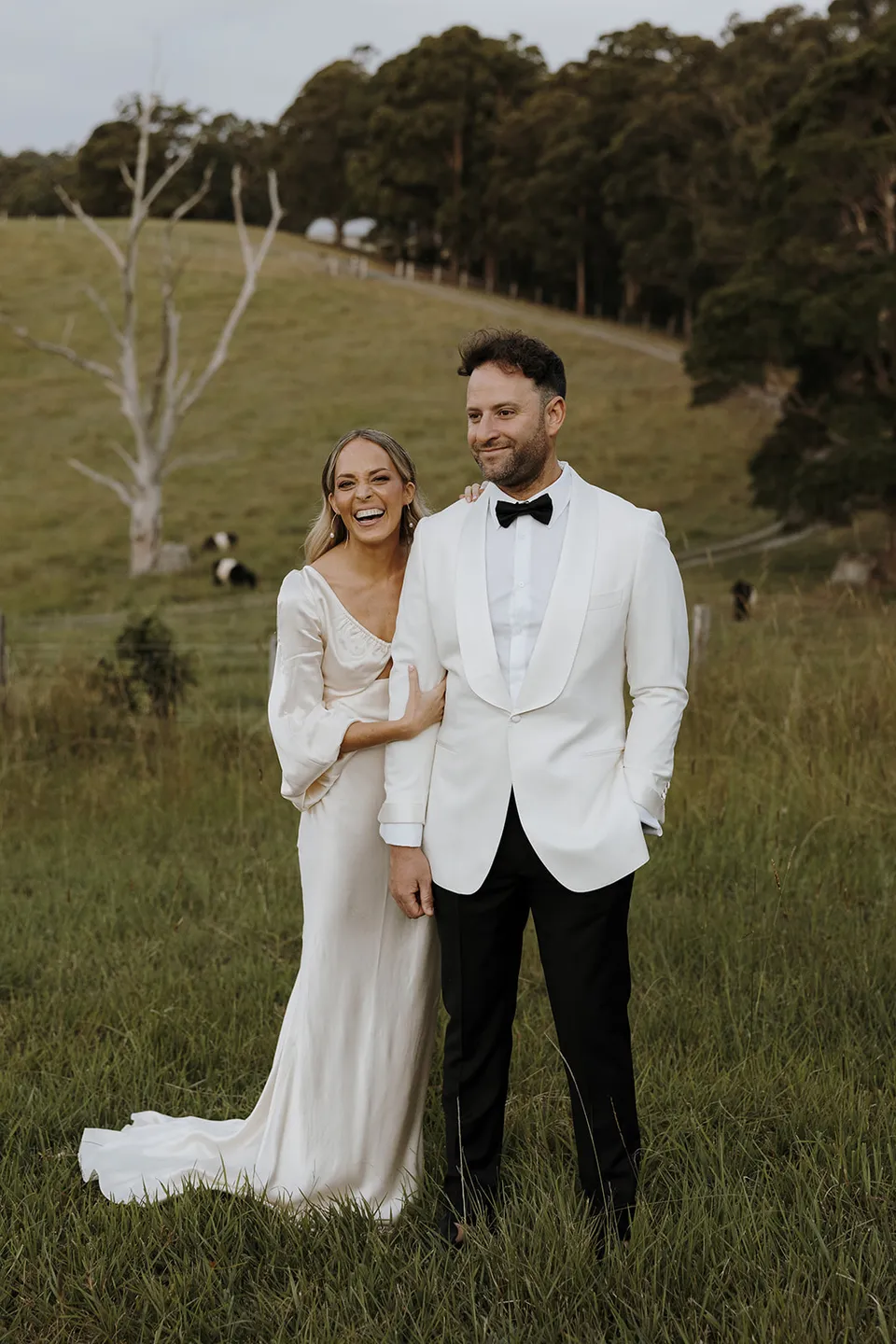 Gabrielle and Greg laughing together in a paddock with green hills and trees behind them, at Roundhill Retreat in Obi Obi, Sunshine Coast Hinterland, Queensland, for an Elopement Collective elopement. Photo by George Bowden.