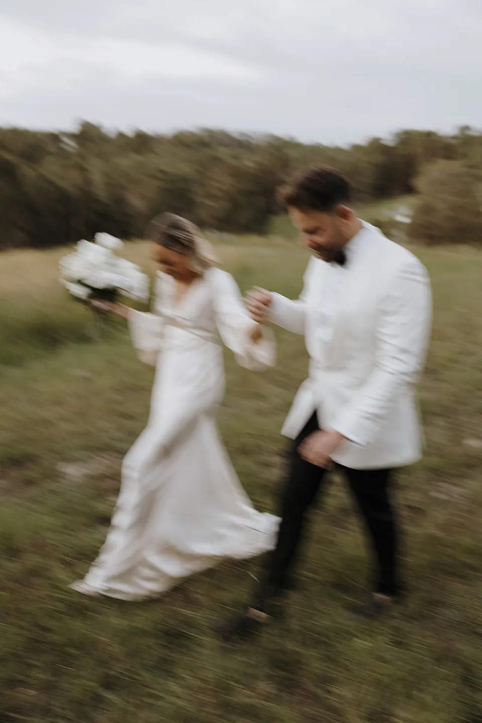 Softly blurred photograph of Gabrielle and Greg walking through the grass, holding hands and her white bouquet by Mrs Gibbons, at Roundhill Retreat in Obi Obi, Sunshine Coast Hinterland, Queensland, for an Elopement Collective elopement. Photo by George Bowden.