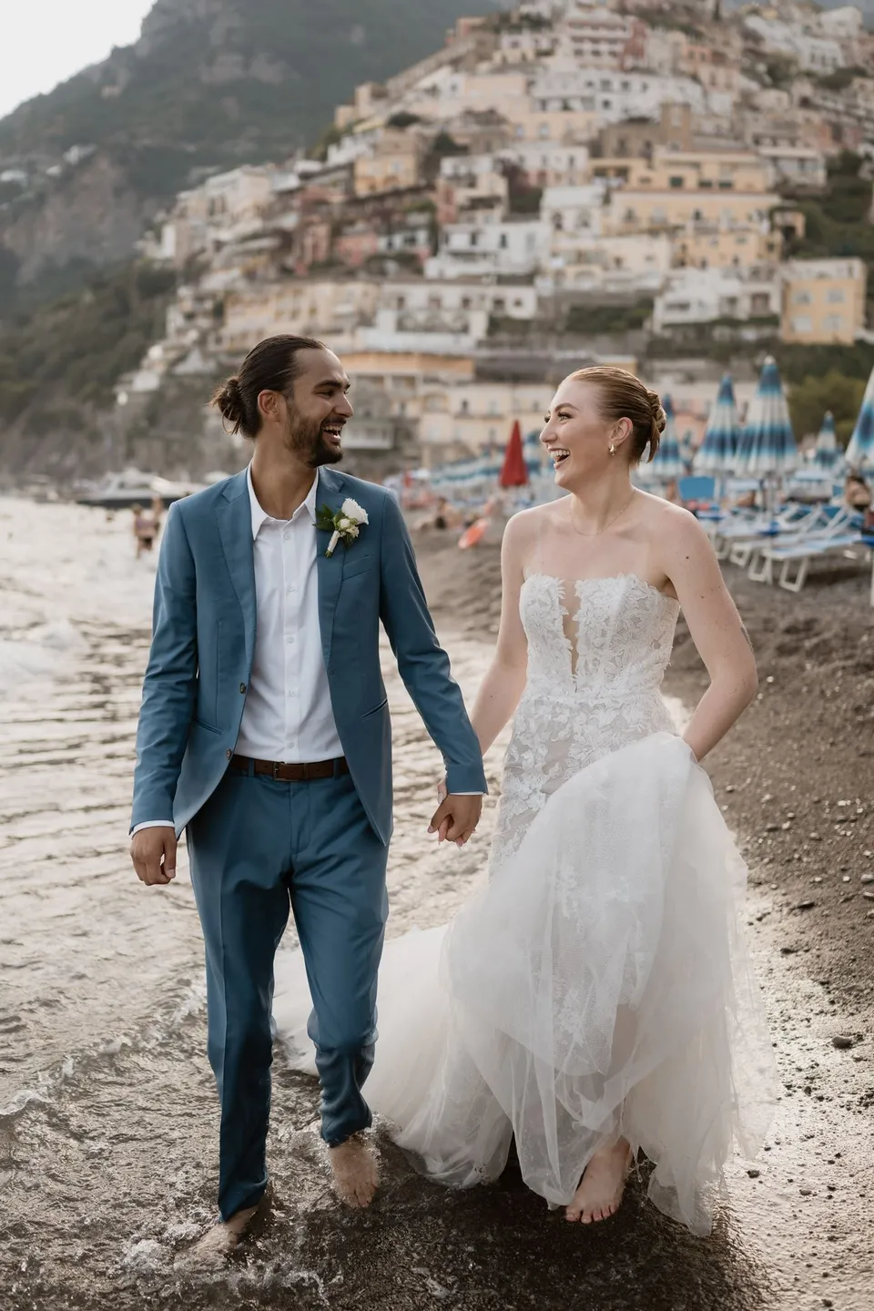 Skyler and Zach laughing together as they walk barefoot in the shallows, holding hands with the hillside town behind them, in Positano on the Amalfi Coast, Italy, for an Elopement Collective elopement. Photograph by Jason Corroto.