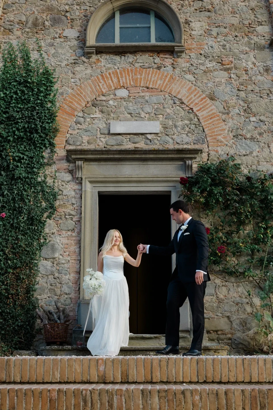 Jack taking Shelby’s hand as she steps through a tall arched stone doorway, in Umbria, Italy, for an Elopement Collective elopement. Photo by James from House of Love.