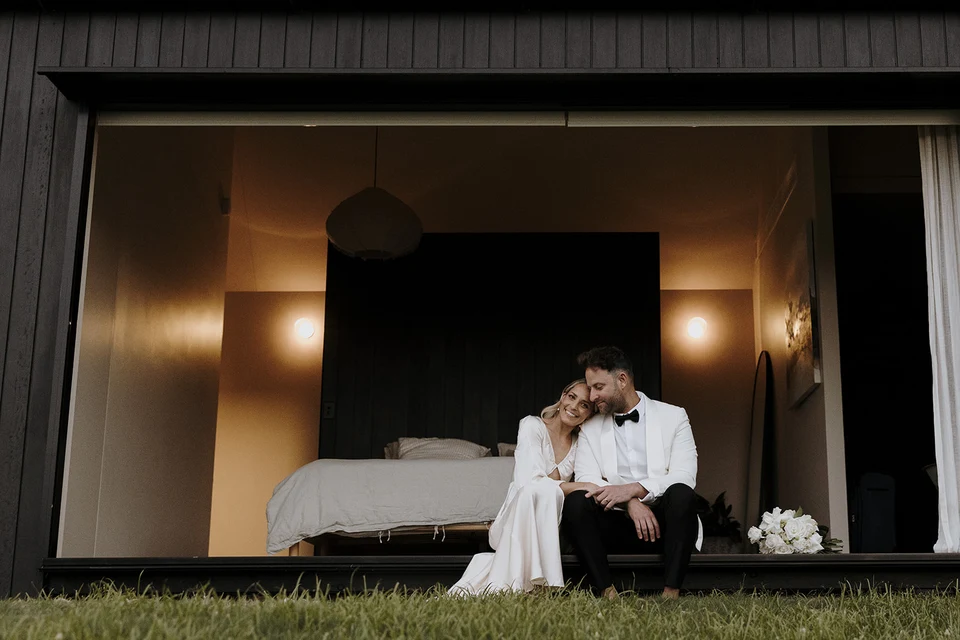 Gabrielle and Greg sitting together in the open bedroom doorway, framed by the cabin’s dark timber exterior, at Roundhill Retreat in Obi Obi, Sunshine Coast Hinterland, Queensland, for an Elopement Collective elopement. Photo by George Bowden.