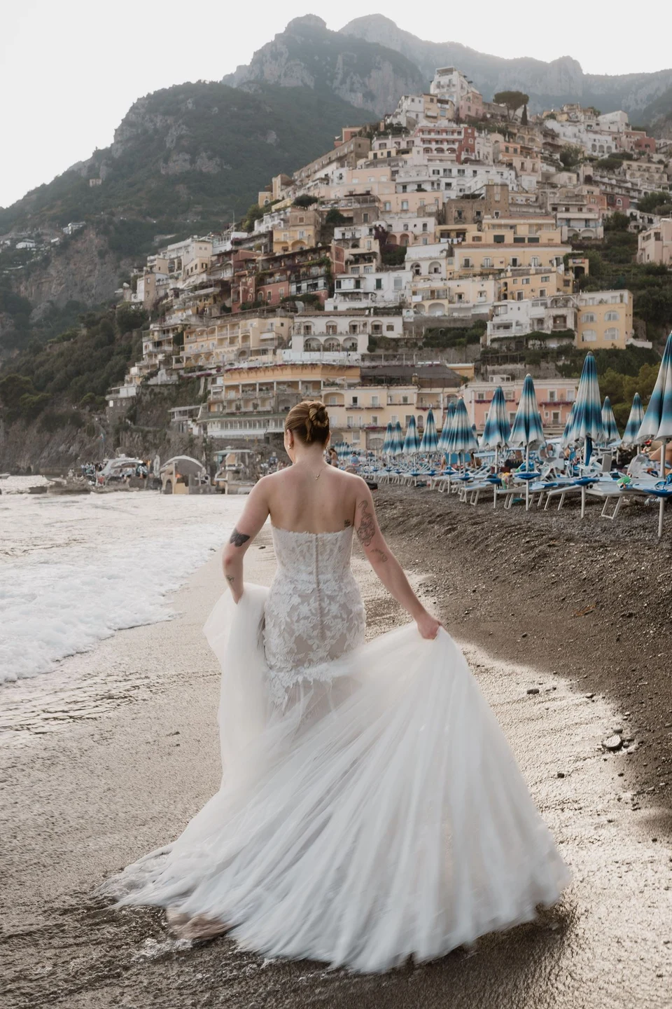 Skyler walking into the shallows with her dress train spread behind her, facing Positano’s hillside houses, in Positano on the Amalfi Coast, Italy, for an Elopement Collective elopement. Photograph by Jason Corroto.