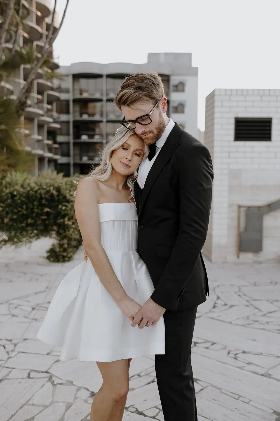 Adam kissing Danielle’s forehead as she leans into him on the stone terrace, at The Calile Hotel in Fortitude Valley, Brisbane, for an Elopement Collective elopement. Photo by George Bowden.