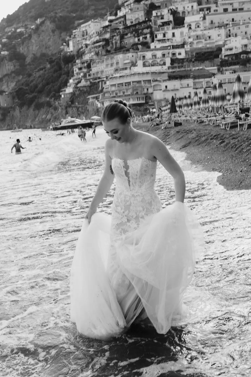 Black and white photograph of Skyler looking down as she gathers her dress and wades in the sea, in Positano on the Amalfi Coast, Italy, for an Elopement Collective elopement. Photograph by Jason Corroto.