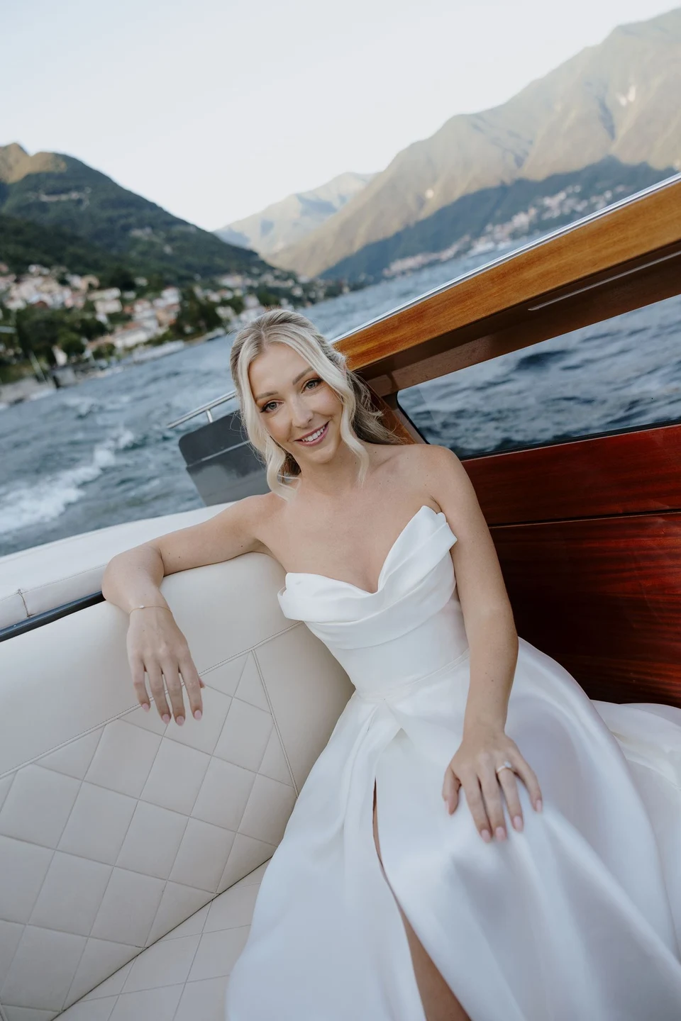 Zoe smiling from a white upholstered seat inside a wooden boat, with lakeside homes and mountains behind her, on Lake Como in Lombardy, Italy, for an Elopement Collective elopement. Photo by George Bowden.