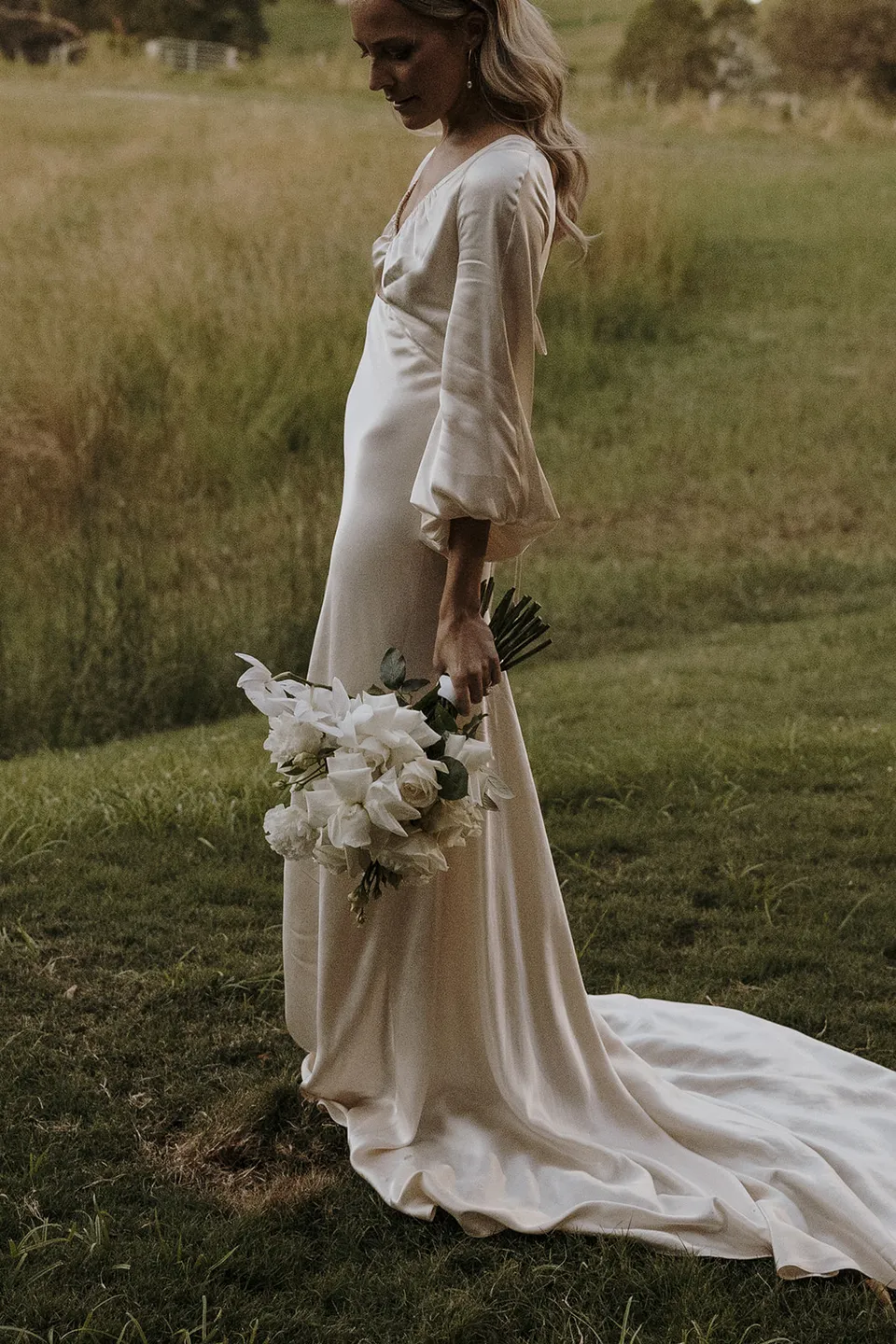 Gabrielle looking down at her white bouquet by Mrs Gibbons in the paddock, her ivory Shona Joy dress trailing through the grass, at Roundhill Retreat in Obi Obi, Sunshine Coast Hinterland, Queensland, for an Elopement Collective elopement. Photo by George Bowden.