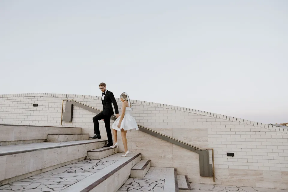 Danielle and Adam climbing wide stone steps bordered by white brick, at The Calile Hotel in Fortitude Valley, Brisbane, for an Elopement Collective elopement. Photo by George Bowden.