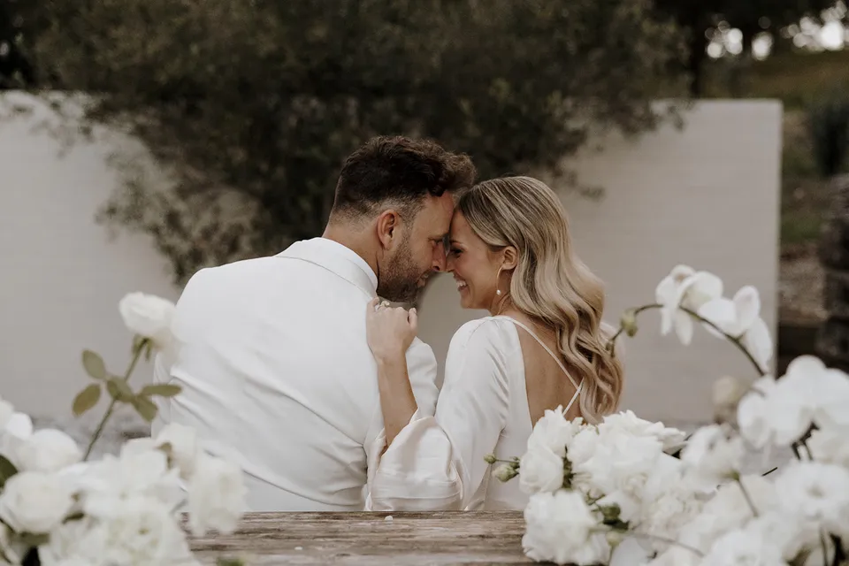 Gabrielle and Greg touching foreheads at a timber courtyard table surrounded by white flowers by Mrs Gibbons, at Roundhill Retreat in Obi Obi, Sunshine Coast Hinterland, Queensland, for an Elopement Collective elopement. Photo by George Bowden.