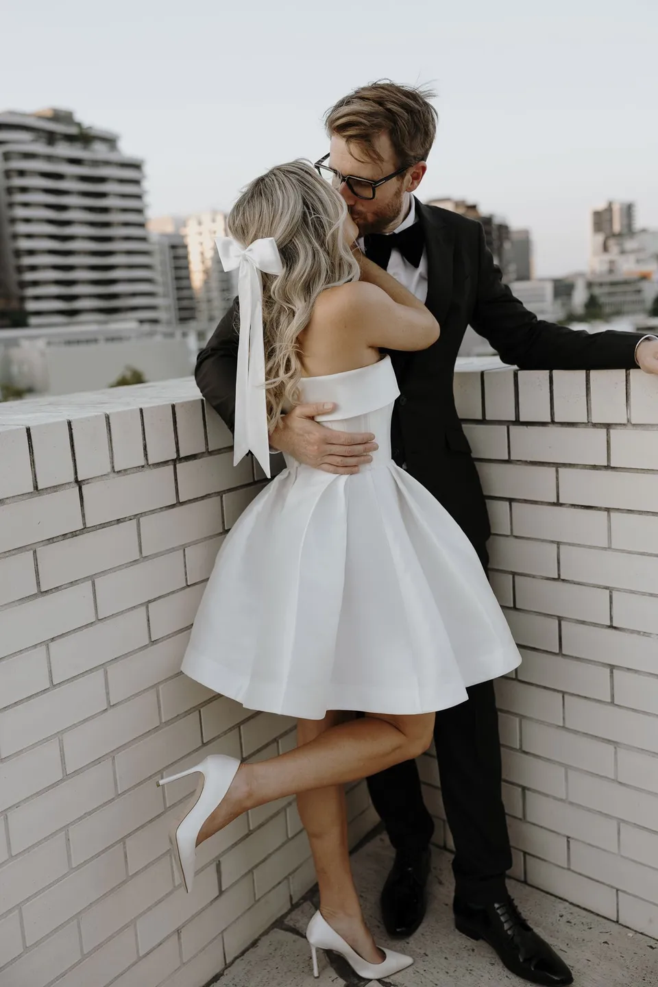 Danielle and Adam embracing beside a white brick balustrade with Brisbane beyond, at The Calile Hotel in Fortitude Valley, Brisbane, for an Elopement Collective elopement. Photo by George Bowden.