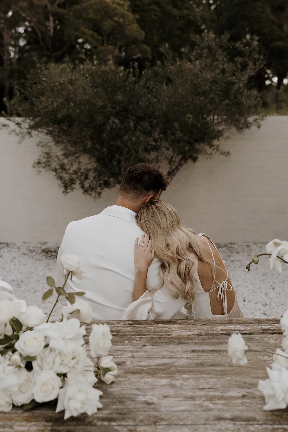 Gabrielle resting her head against Greg at the courtyard table, with white flowers by Mrs Gibbons in the foreground, at Roundhill Retreat in Obi Obi, Sunshine Coast Hinterland, Queensland, for an Elopement Collective elopement. Photo by George Bowden.