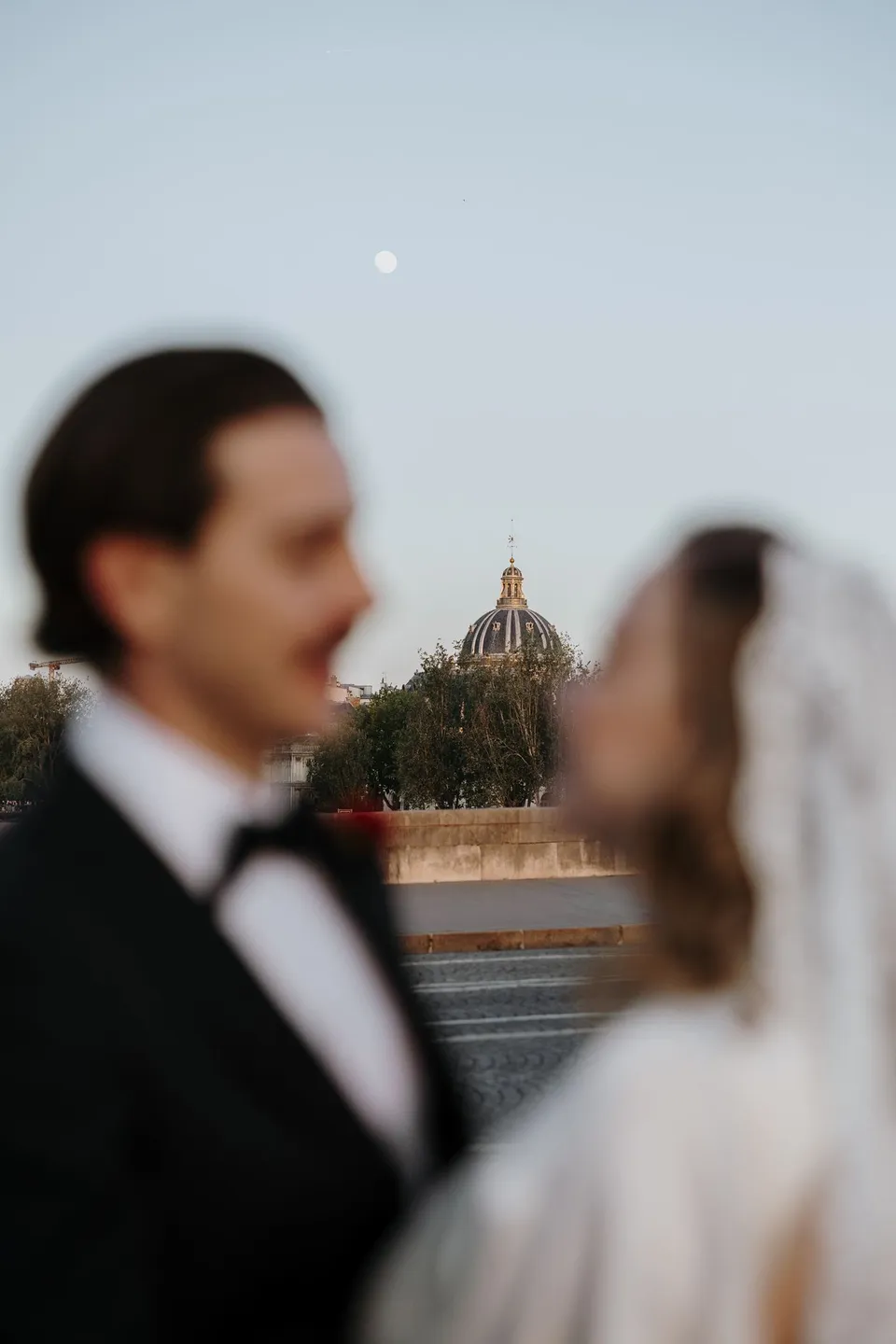 A soft-focus portrait of Kara and Samuel facing each other, with a small moon and distant Parisian rooftops behind them, beside the Seine in Paris, France, during an Elopement Collective elopement. Photograph by George Bowden.