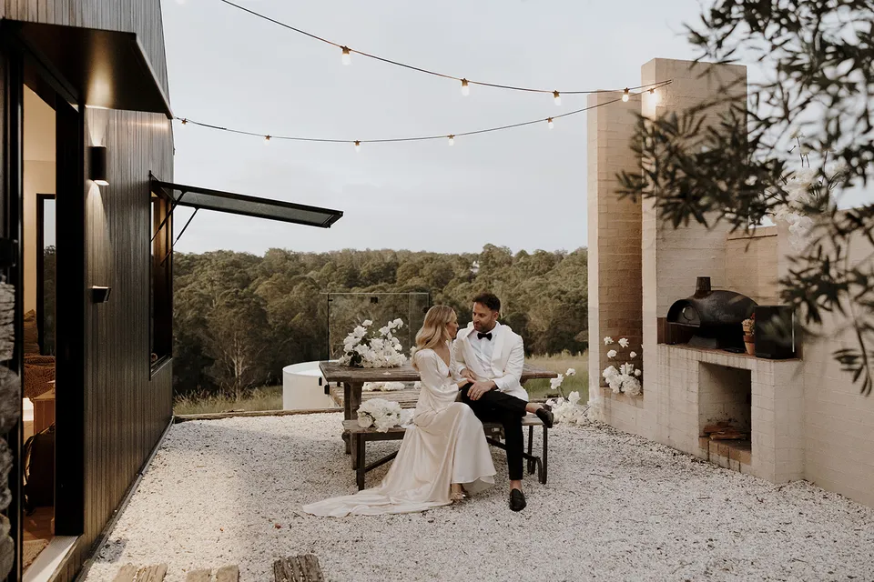 Gabrielle and Greg sitting together in the cabin courtyard beneath festoon lights, with the outdoor fireplace beside them, at Roundhill Retreat in Obi Obi, Sunshine Coast Hinterland, Queensland, for an Elopement Collective elopement. Photo by George Bowden.