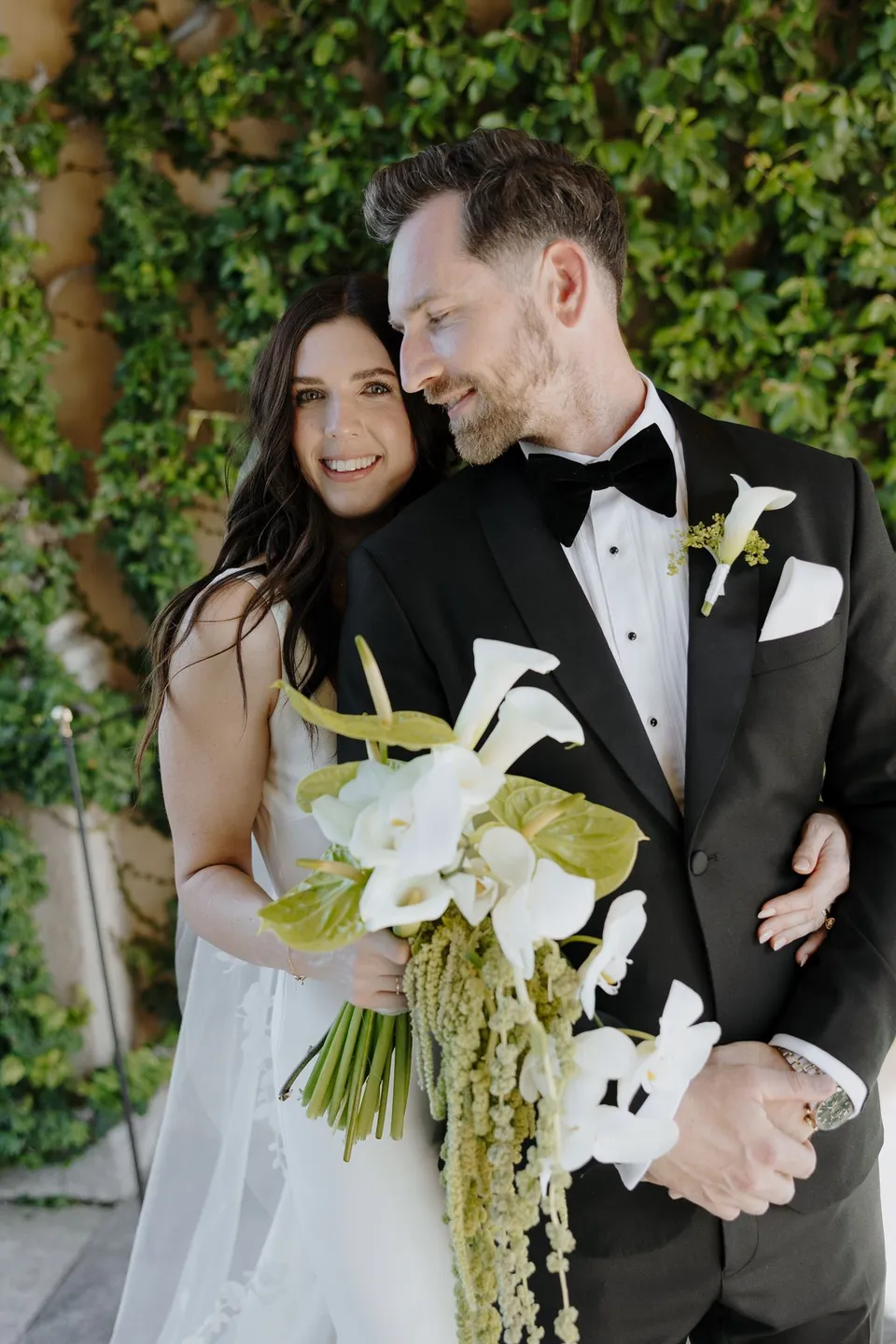 Lyndal and Lachlan smiling together beside an ivy-covered wall at Villa del Balbianello, with a white bouquet and trailing greenery by Garden of Love, at Lake Como in Lombardy, Italy, for an Elopement Collective elopement. Photograph by George Bowden.
