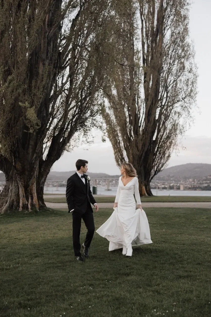 A couple walk across a lawn beneath a line of tall trees.