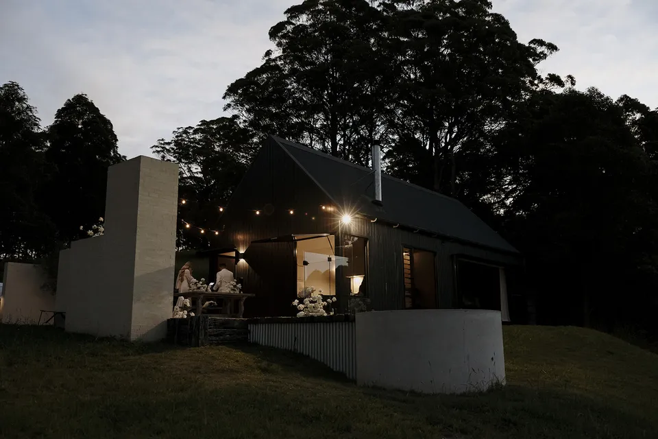 The cabin glowing beneath festoon lights at dusk, with white flowers by Mrs Gibbons in the courtyard, at Roundhill Retreat in Obi Obi, Sunshine Coast Hinterland, Queensland, for an Elopement Collective elopement. Photo by George Bowden.