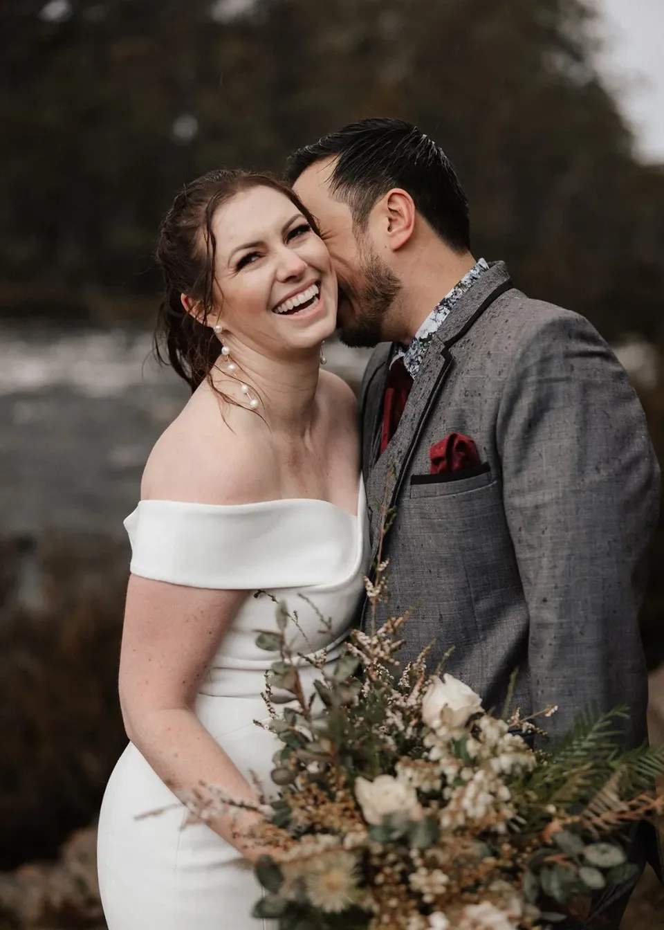 Sarah laughing with her bouquet by The Romantics in hand as Jono leans close to her cheek, at Pumphouse Point, Lake St Clair, Tasmania, for an Elopement Collective elopement. Photo by Bulb Creative.