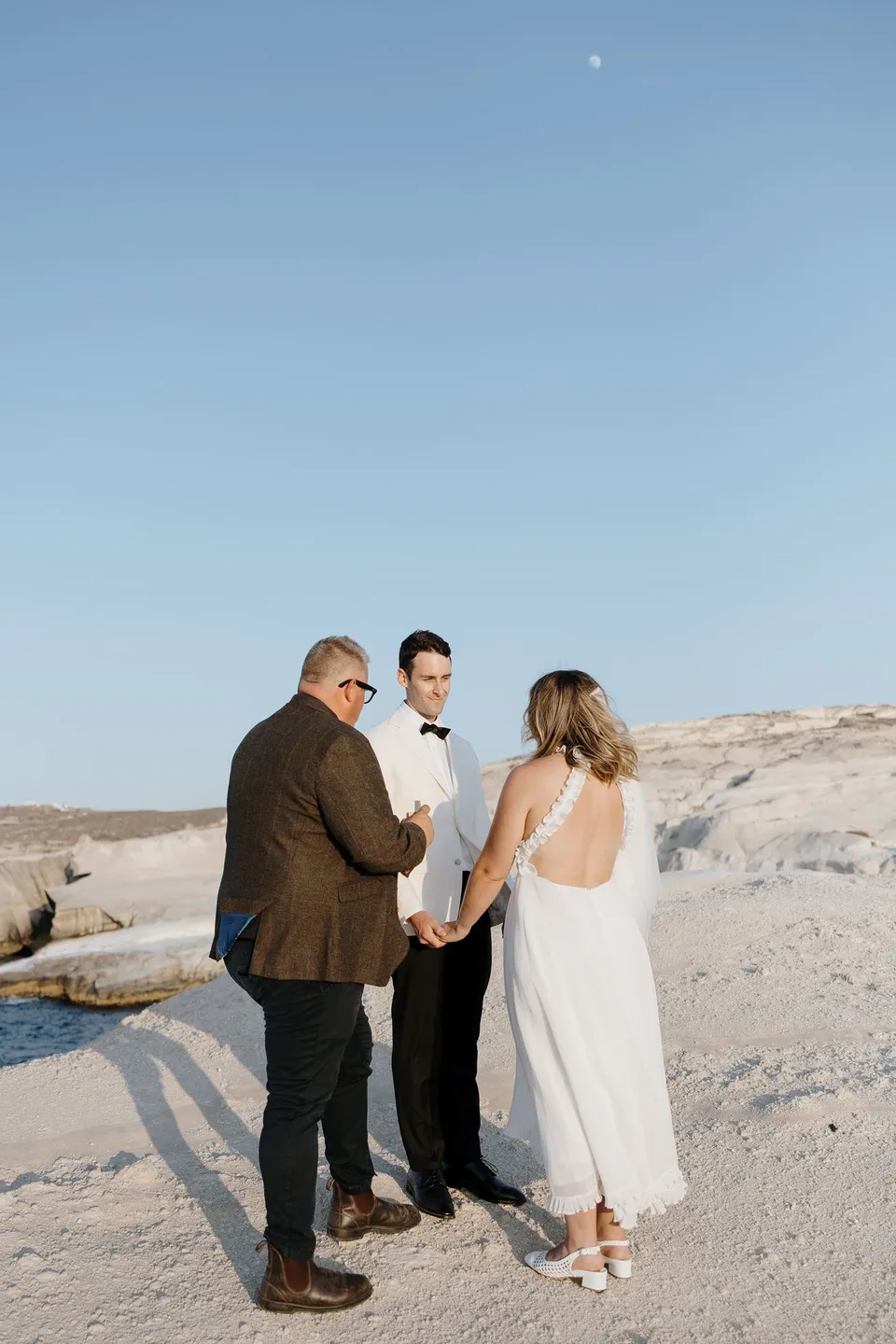 Celebrant Josh Withers speaking as Georgina and Brad hold hands during their ceremony, with the sea and white rocks behind them, at Sarakiniko on Milos in the Cyclades, Greece, during an Elopement Collective elopement. Photograph by George Bowden.