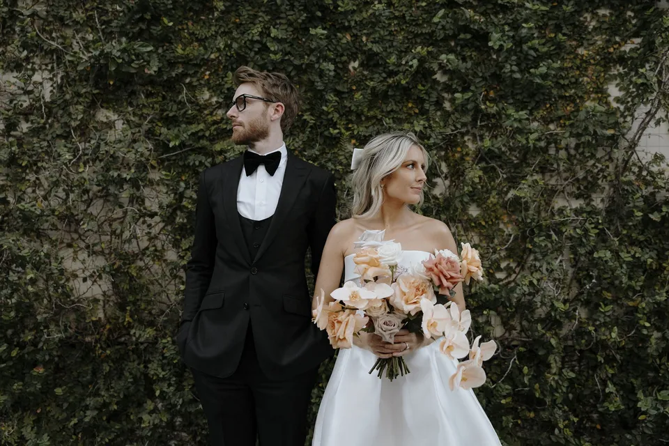 Danielle holding her peach and white bouquet by Mrs Gibbons Florals beside Adam in front of a leafy ivy wall, in the James Street precinct, Fortitude Valley, Brisbane, for an Elopement Collective elopement. Photo by George Bowden.