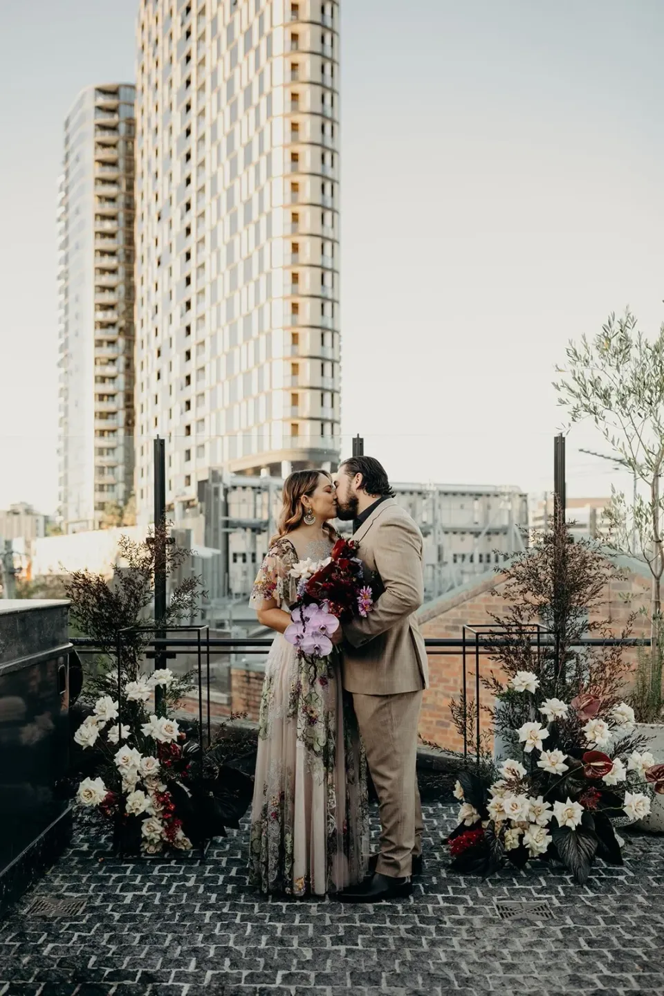 Victoria and Daniel embracing among rooftop ceremony flowers by Mrs Gibbons Flowers, at Agnes Restaurant in Fortitude Valley, Brisbane, for an Elopement Collective elopement. Photo by Tessa Shannon.