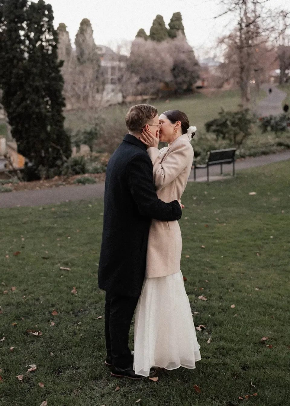 A couple kiss on a garden lawn with tall trees and historic buildings beyond.