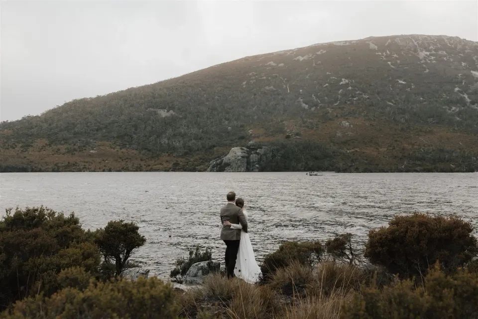 A couple embrace at the edge of a mountain lake surrounded by low shrubs.