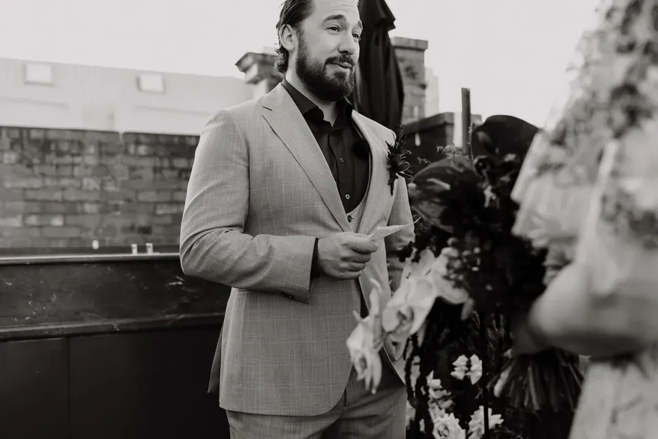 Black-and-white photograph of Daniel reading his vows to Victoria on the rooftop terrace, at Agnes Restaurant in Fortitude Valley, Brisbane, for an Elopement Collective elopement. Photo by Tessa Shannon.