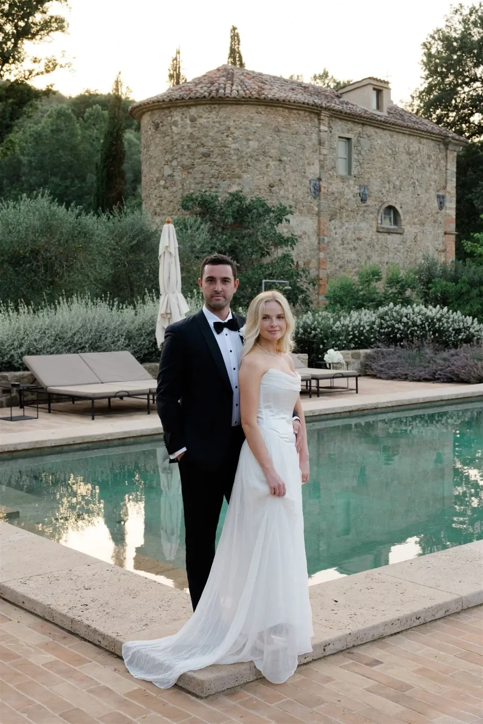 Shelby and Jack standing together beside a turquoise swimming pool and garden terrace, in Umbria, Italy, for an Elopement Collective elopement. Photo by James from House of Love.
