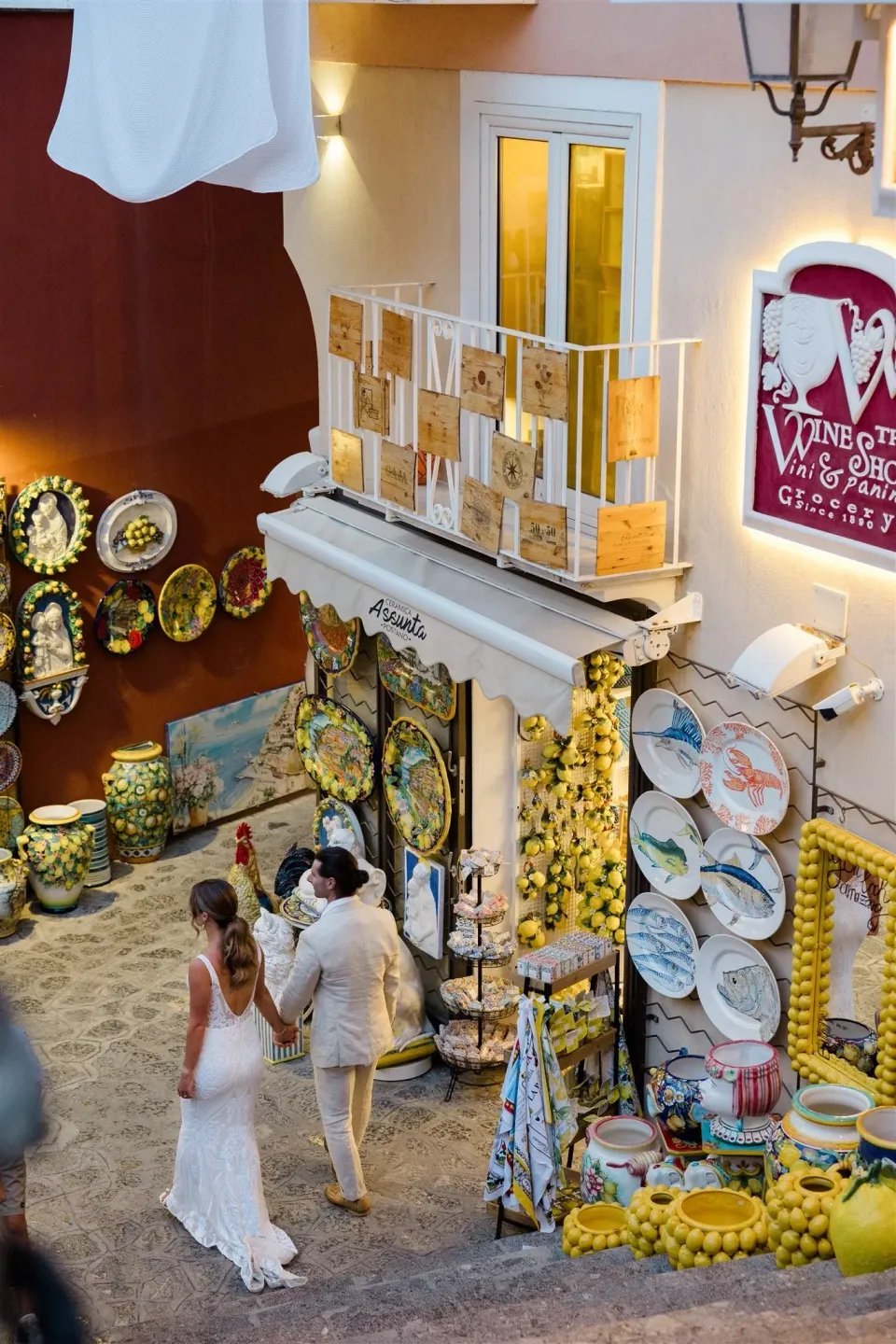 Sarah and Josh walking hand in hand past a colourful ceramics shop on a narrow staircase, in Positano on Italy’s Amalfi Coast, for an Elopement Collective elopement. Photo by James from House of Love.