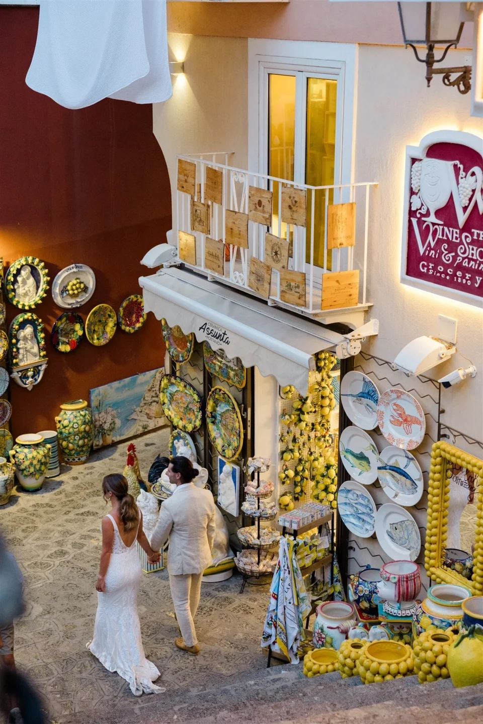 Sarah and Josh walking hand in hand past a colourful ceramics shop on a narrow staircase, in Positano on Italy’s Amalfi Coast, for an Elopement Collective elopement. Photo by James from House of Love.