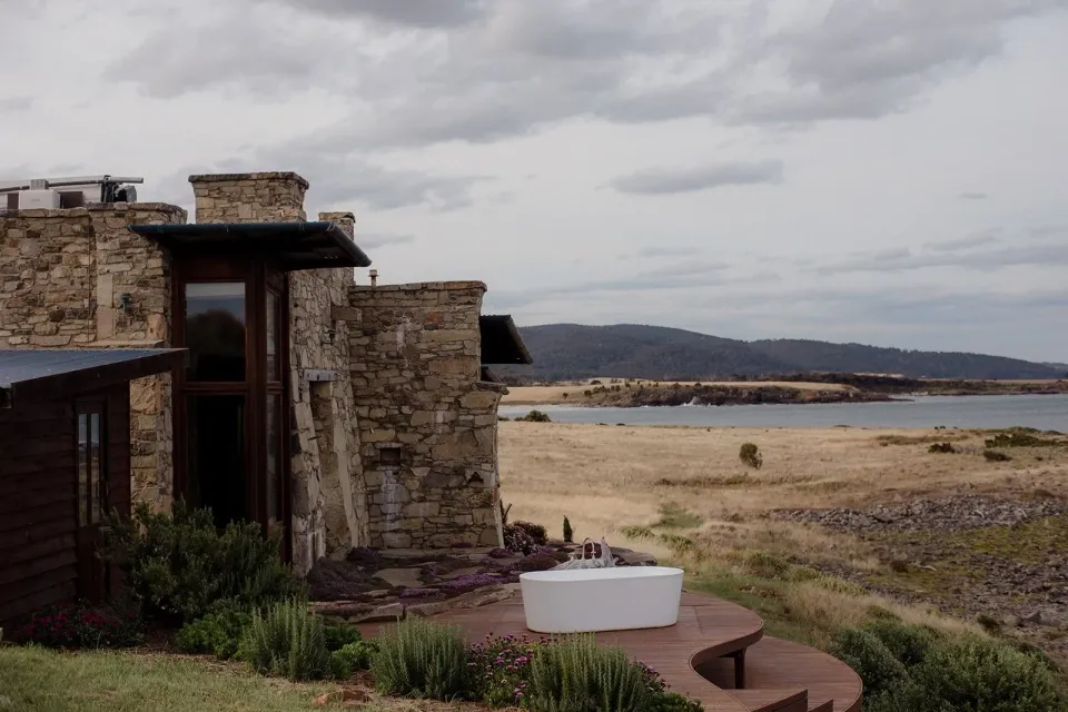 A stone cottage with an outdoor bath overlooks coastal grass and the sea.