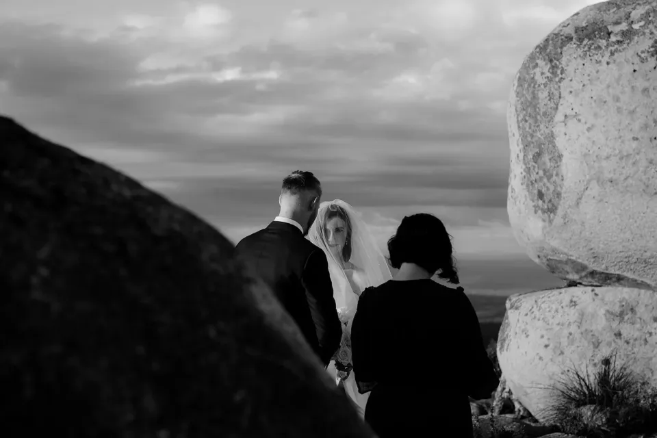 A black-and-white photograph of a couple and celebrant among large coastal boulders.