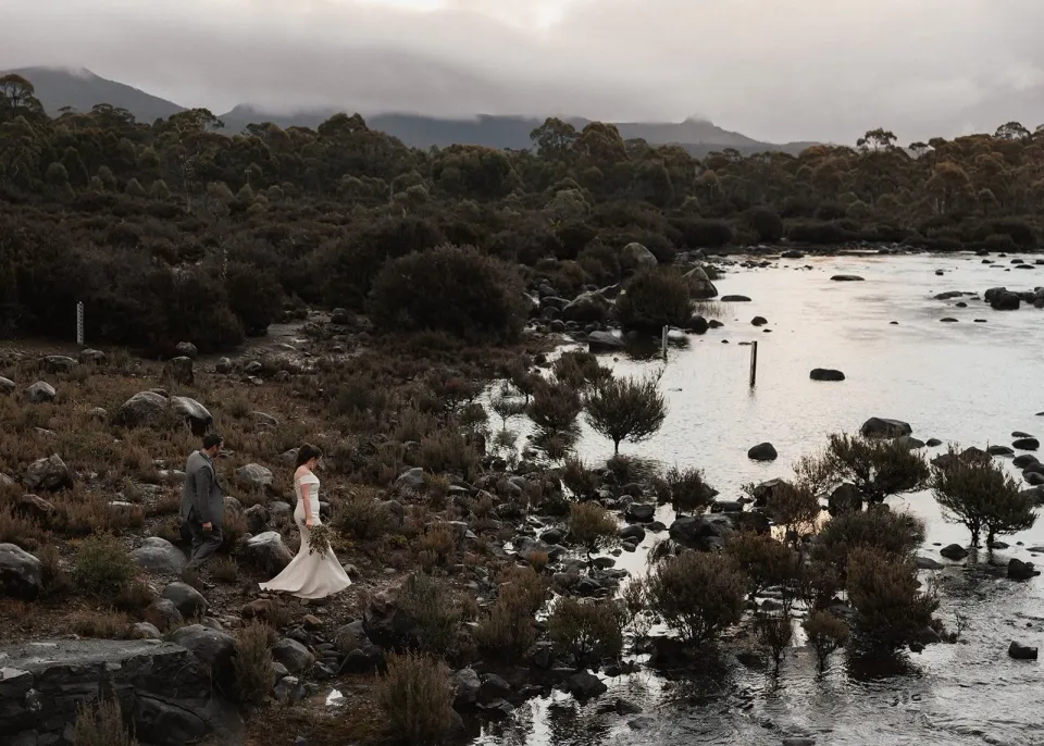 A couple stand beside a lake edged with rocks, shrubs and forest.