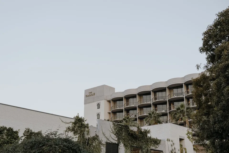 The hotel’s curved balconies and signed white brick exterior above leafy trees, at The Calile Hotel in Fortitude Valley, Brisbane, for an Elopement Collective elopement. Photo by George Bowden.