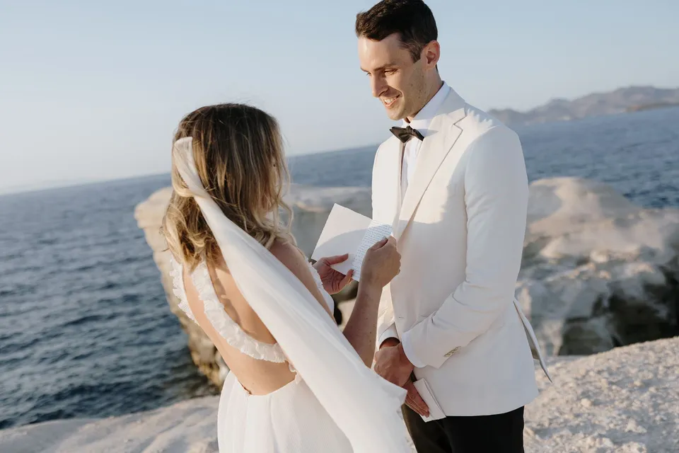 Georgina reading her handwritten personal vows from an open book as Brad smiles and holds her hand, with her veil moving in the breeze, at Sarakiniko on Milos in the Cyclades, Greece, during an Elopement Collective elopement. Photograph by George Bowden.